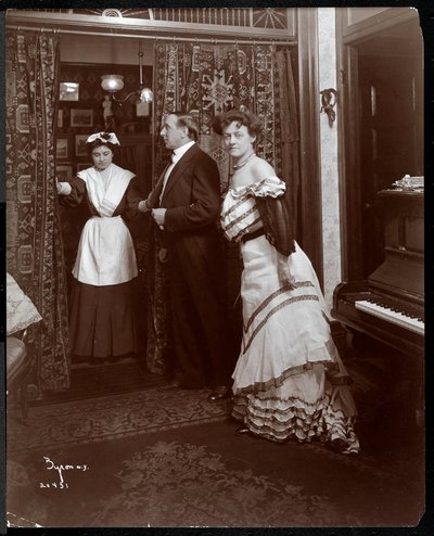 Un uomo e una donna vestiti formalmente e quello che sembra essere un domestico in una stanza con un pianoforte, 1905 da Byron Company